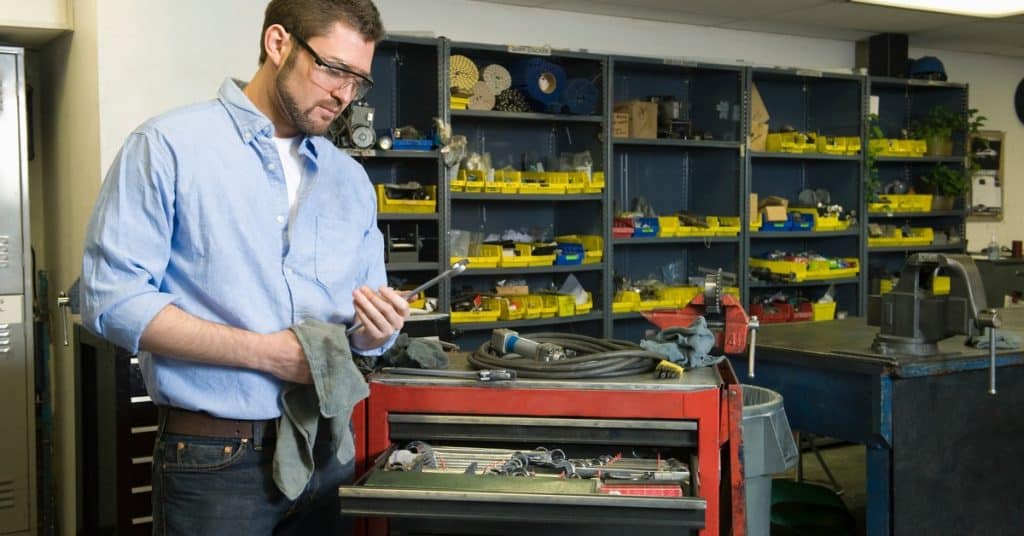 A person wears protective glasses while standing in a workshop near a tool cart. They wipe a tool with a gray cloth.
