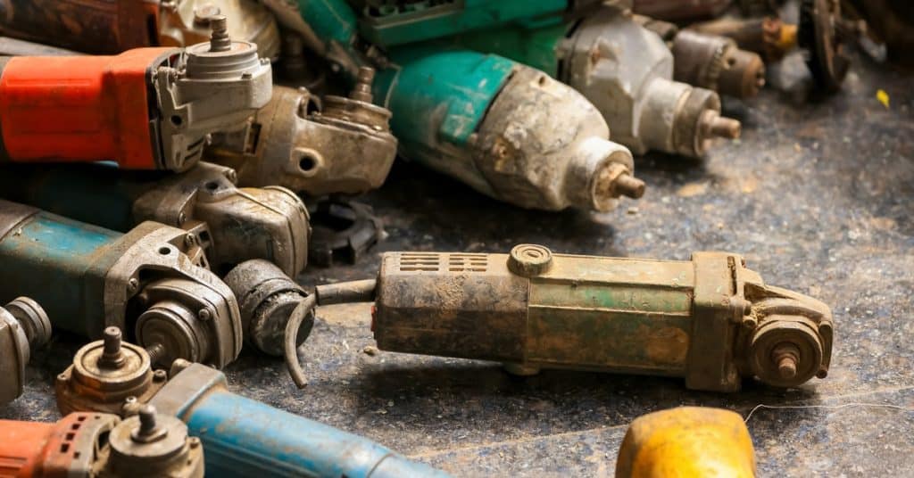 A pile of rusty power tools lies on the ground. Some tools have red, blue, or yellow components.