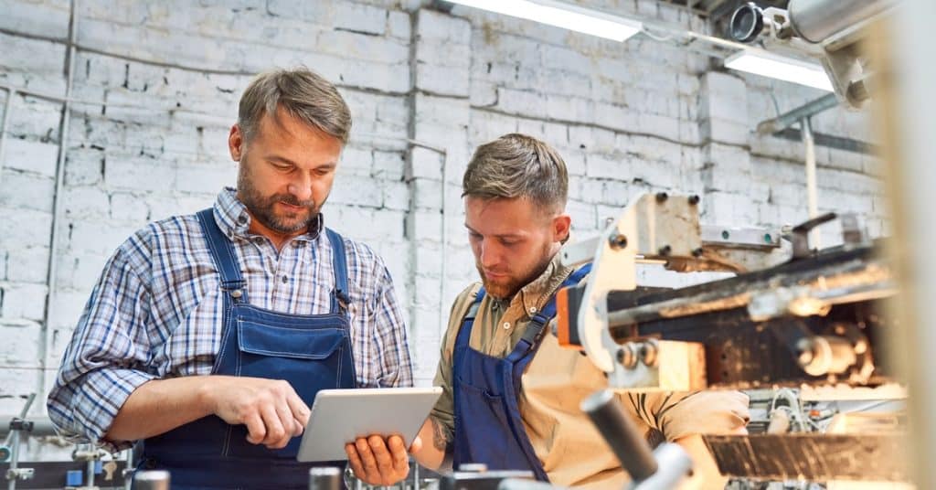 Two men stand beside each other in a machine shop. One man holds a gray tablet, and they both look at the screen.