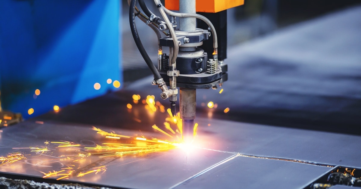A close-up of a machine cutting metal while sparks fly near the incision lines. The background is blurred.