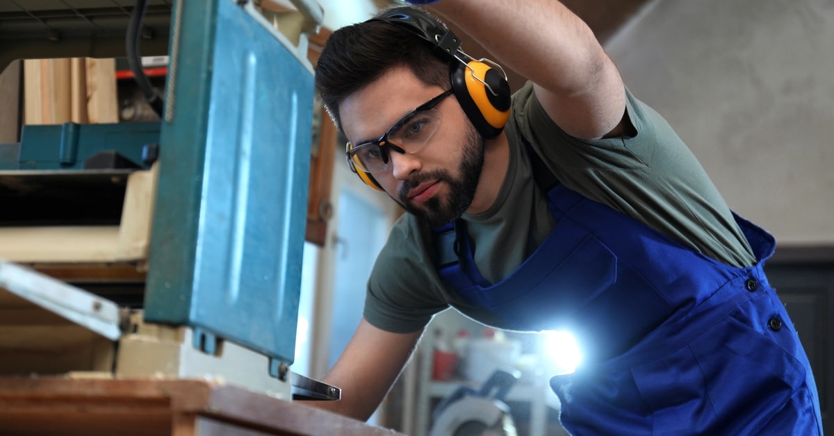 A man is using a machine in a shop. He wears protective eyeglasses, gloves, and a blue work apron.