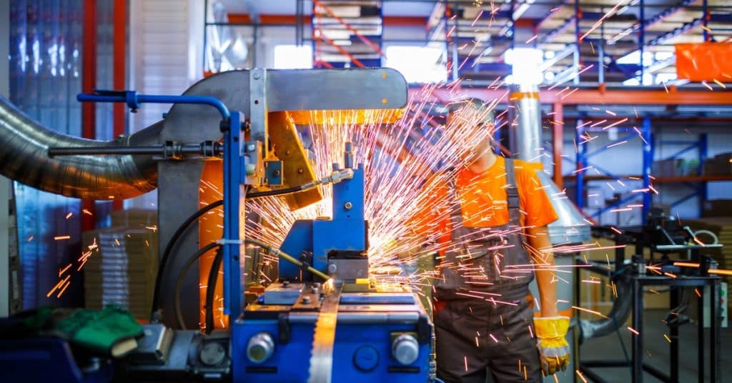 A man stands behind a large welding machine in a machine shop. Sparks fly out of the machine, and it hides the man's face.