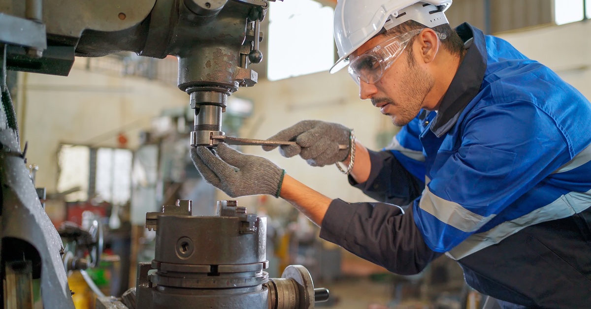 A man using a wrench screws a component to a machine. He wears protective gear, including a hard hat and glasses.