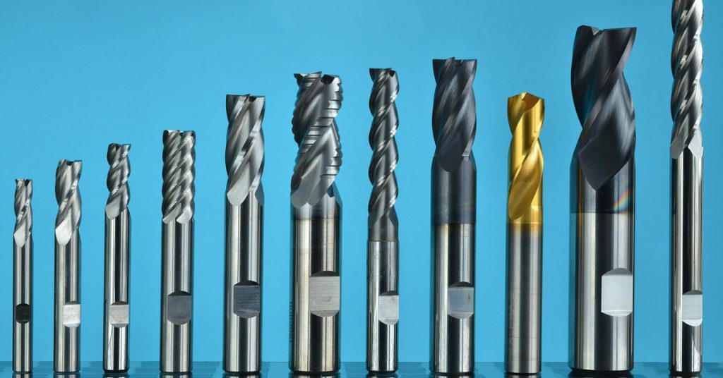A row of drill bits stands on a table in front of a blue wall. Some drill bits have different coatings.