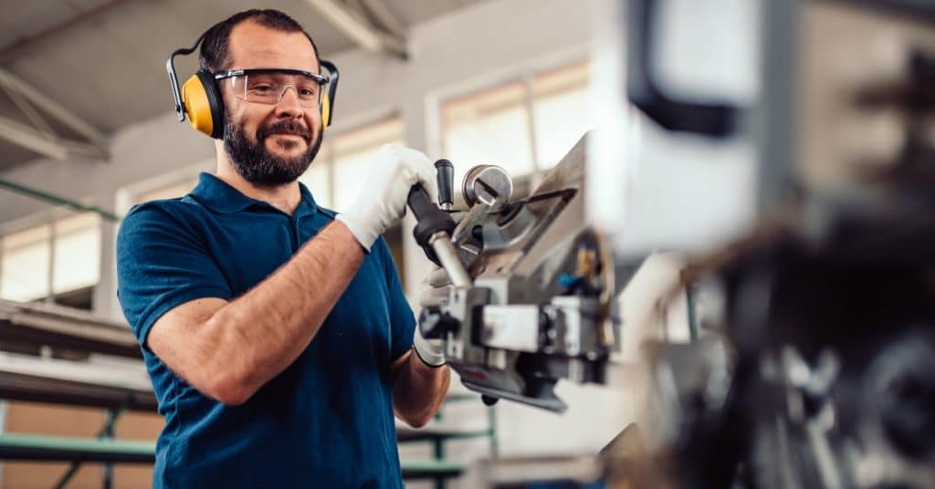 A man operates a machine while wearing ear protection, protective glasses, and gloves. He's also smiling.