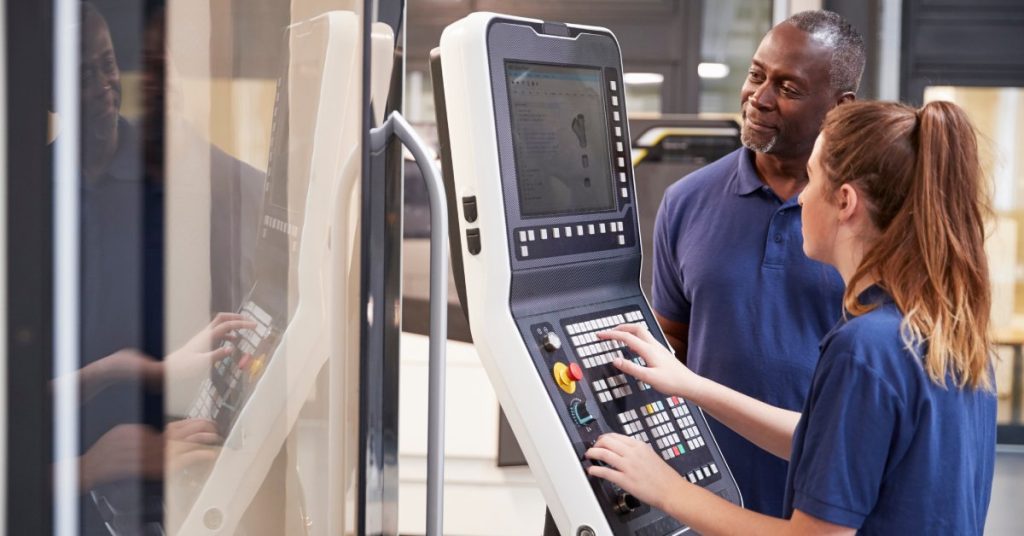 An African American man and a white woman standing near a CNC machine. The woman presses the machine's buttons.