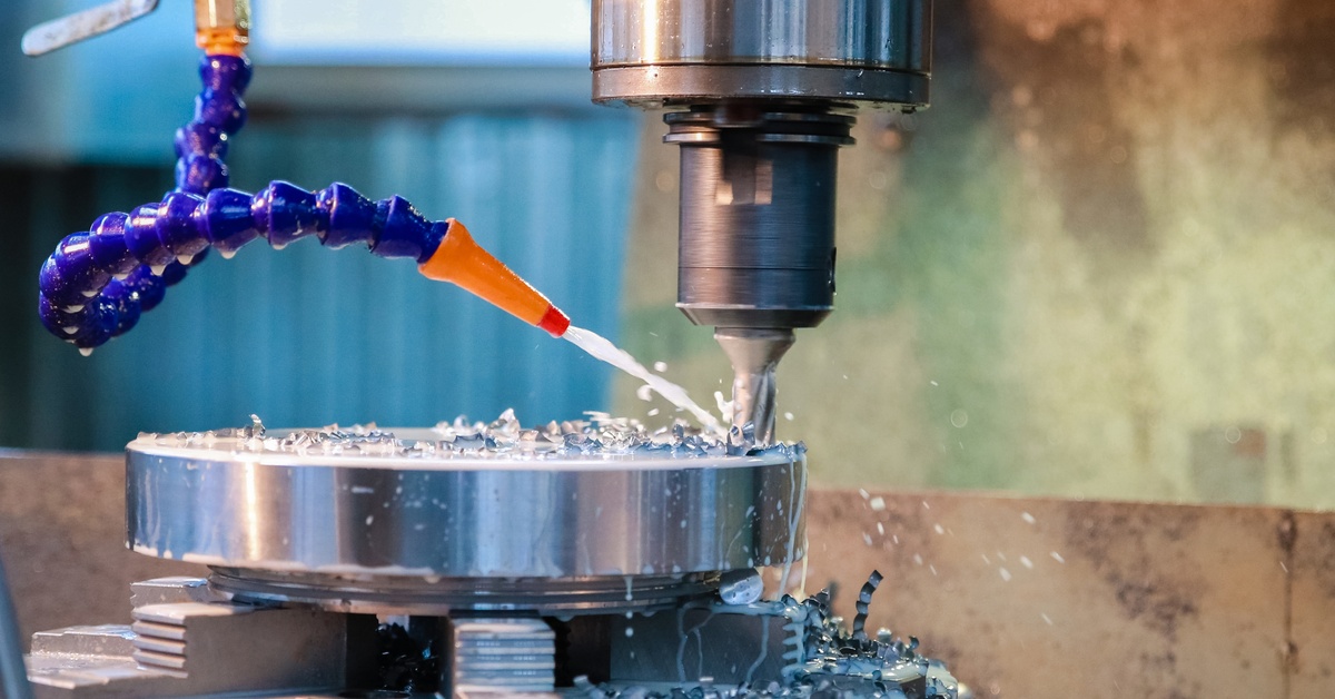 Lubricant from a blue and orange nozzle sprays on a drilling machine. The machine has metal bits on the surface.