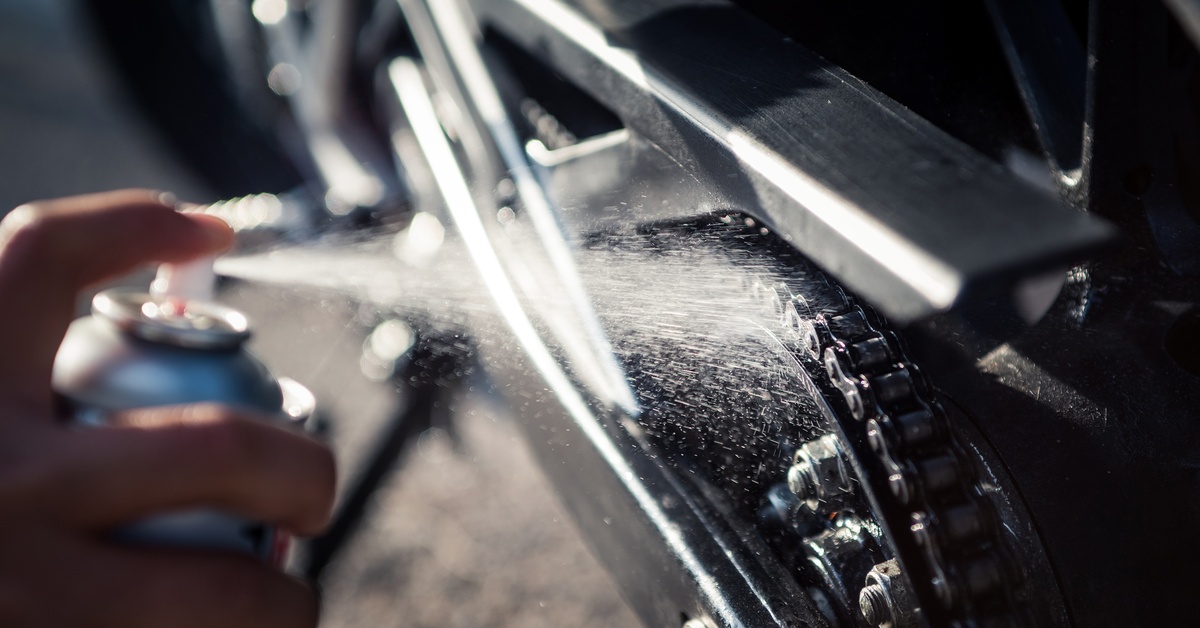 A close-up of someone spraying a water-based lubricant on a machine chain. The machine is slightly blurry.